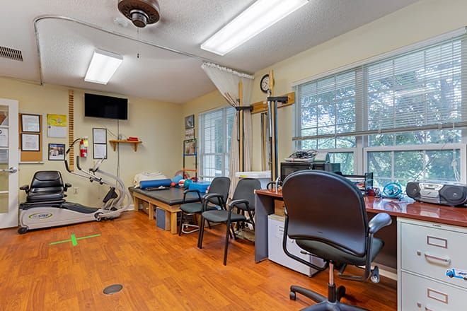 A therapy room with exercise equipment, chairs, desks, and large windows.