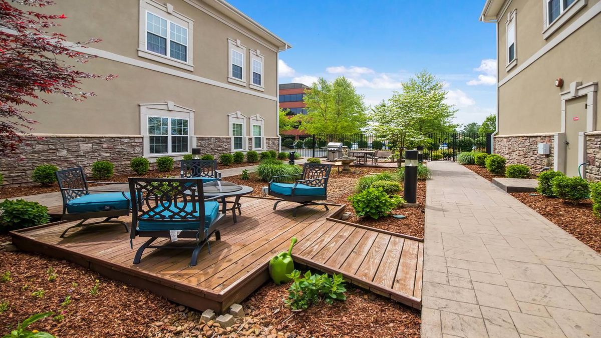 A modern outdoor seating area with a wooden deck surrounded by greenery and adjacent to a building.
