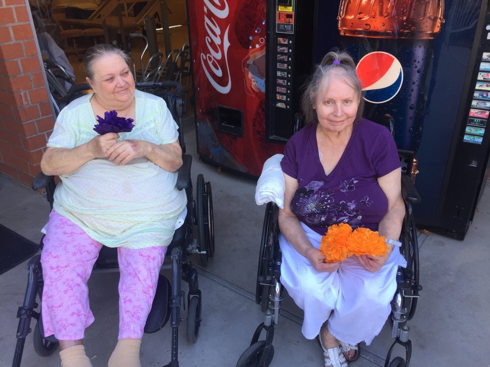 Two elderly women in wheelchairs holding colorful flowers sit outside near vending machines.