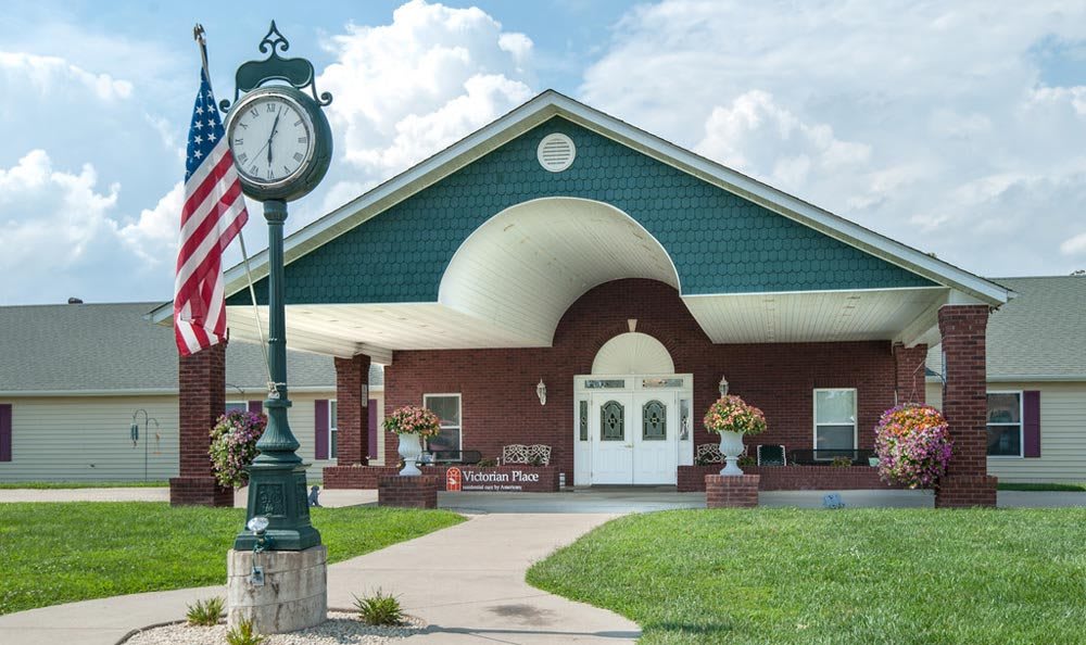 A quaint building with a clock and an American flag stands amidst a lush green lawn under a partly cloudy sky.