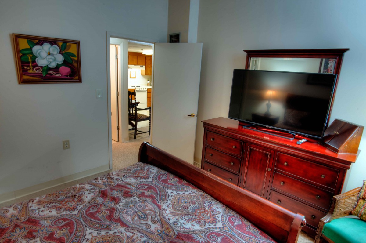 A cozy bedroom featuring a large bed, wooden dresser with a TV on top, and an open door leading to another room.