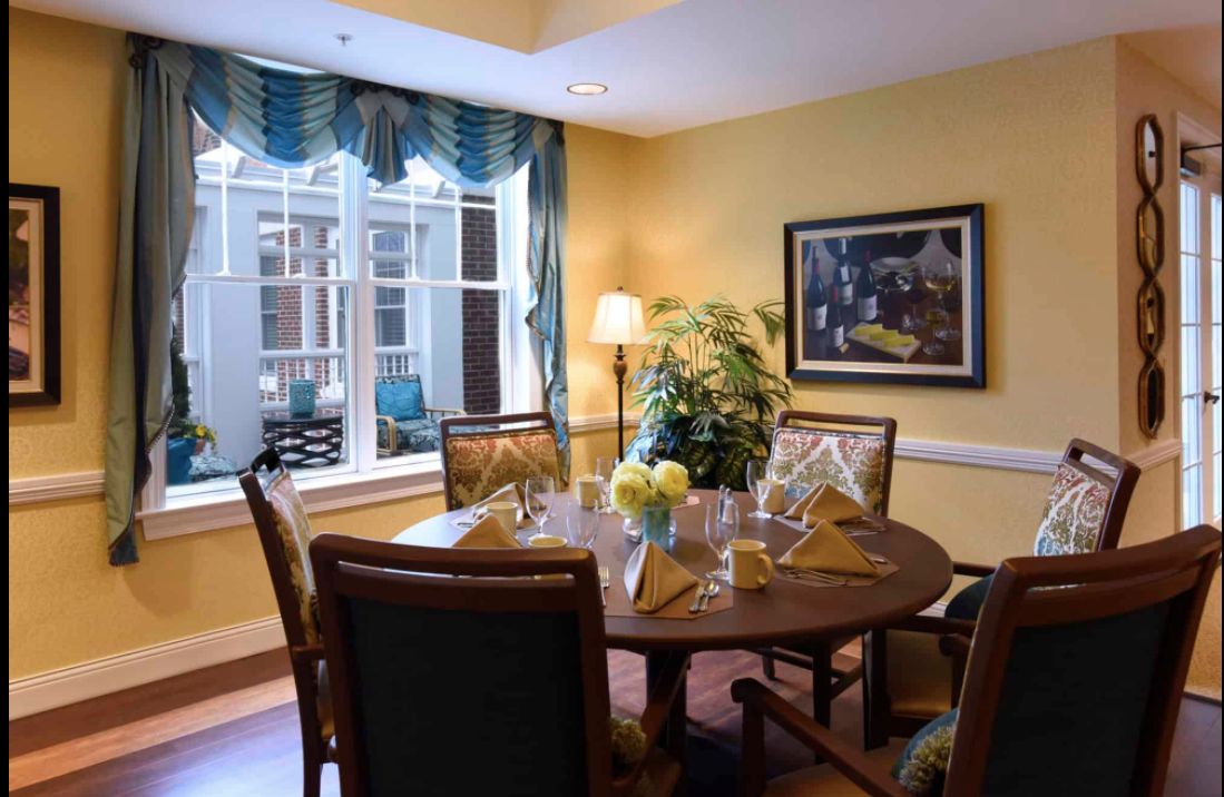 A cozy dining room with a round table set for four, featuring elegant decor and large windows allowing natural light in.