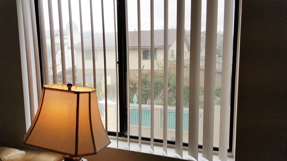 A room with a lamp next to a window covered by vertical blinds, revealing an outdoor scene.