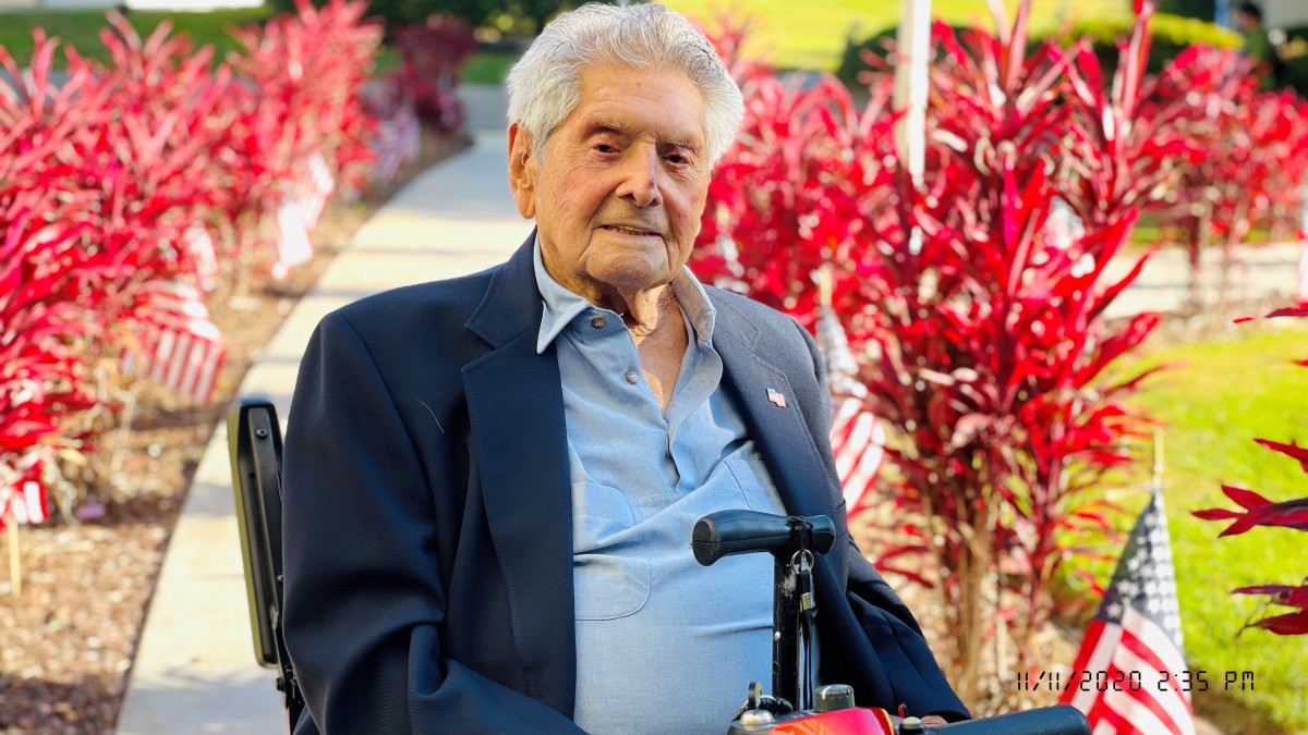 An elderly man in a suit is sitting outdoors in a wheelchair, surrounded by vibrant red plants.