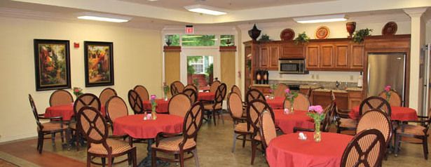 A cozy dining area with round tables covered in red tablecloths and elegant wooden chairs, featuring a kitchenette in the background.