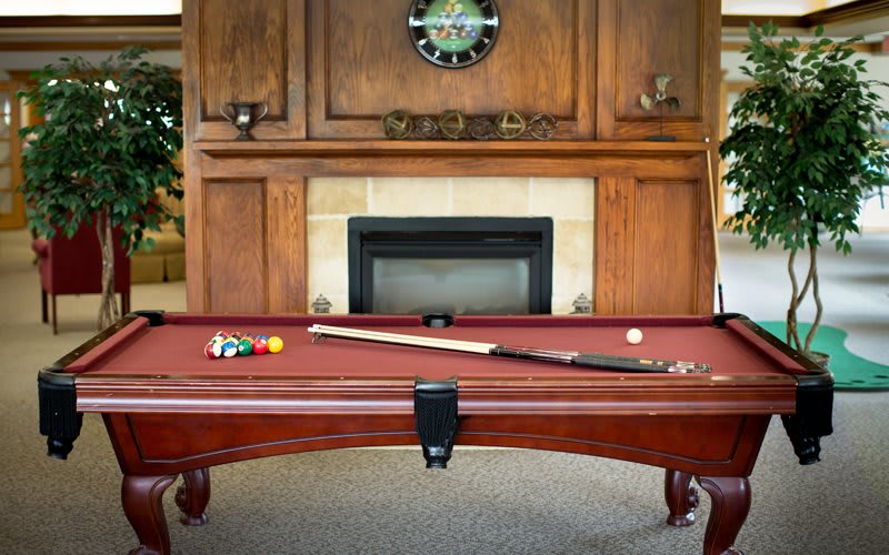 A cozy recreation room featuring a pool table with billiard balls and a cue stick, set against a wooden-paneled fireplace.