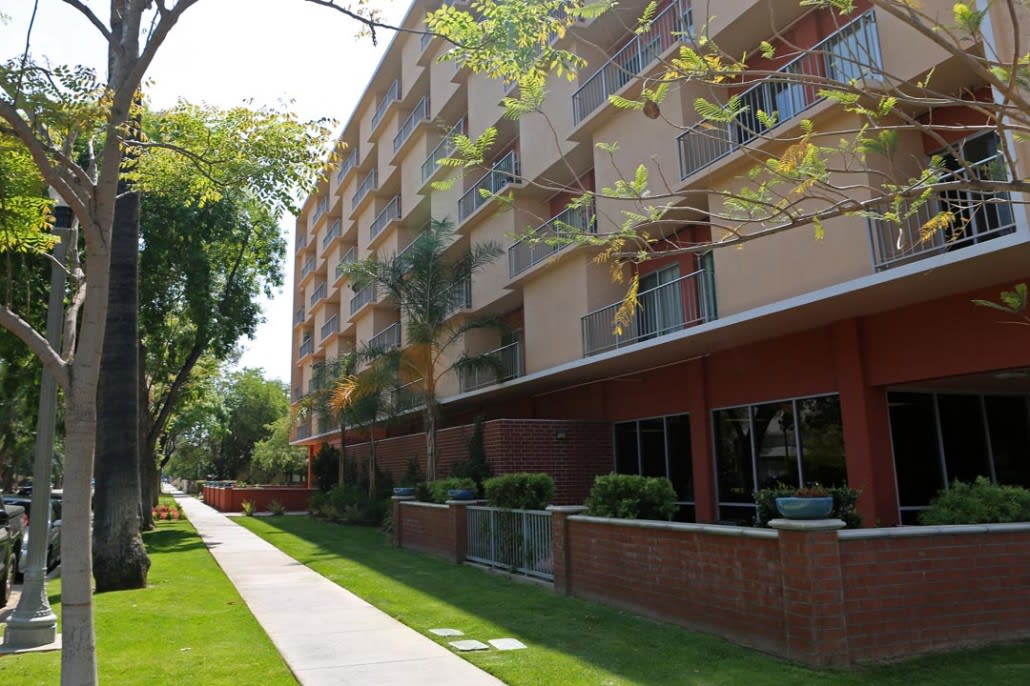 A residential apartment building with balconies, surrounded by greenery and a sidewalk.
