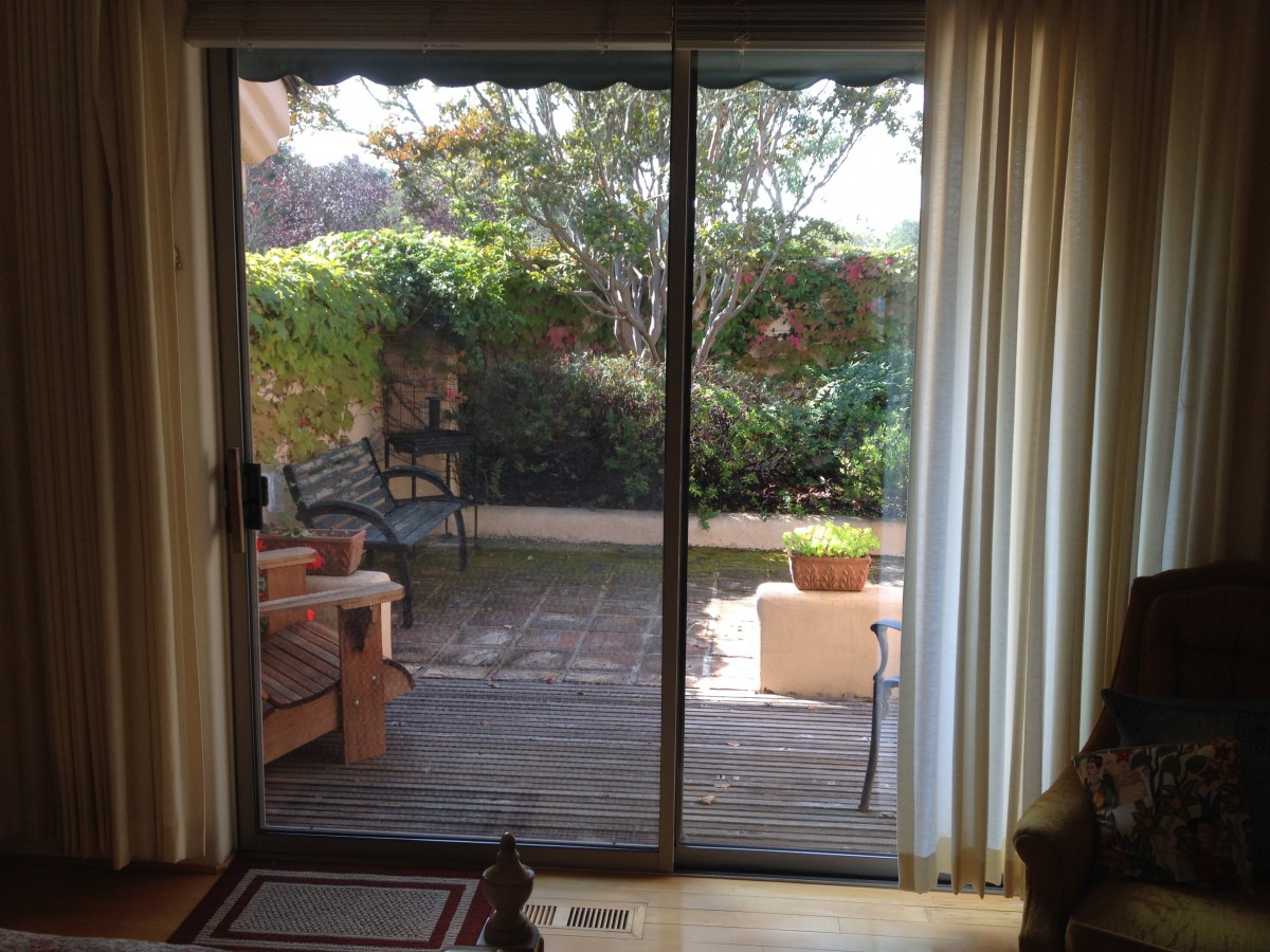 A view of a garden patio through sliding glass doors with outdoor seating and lush greenery.