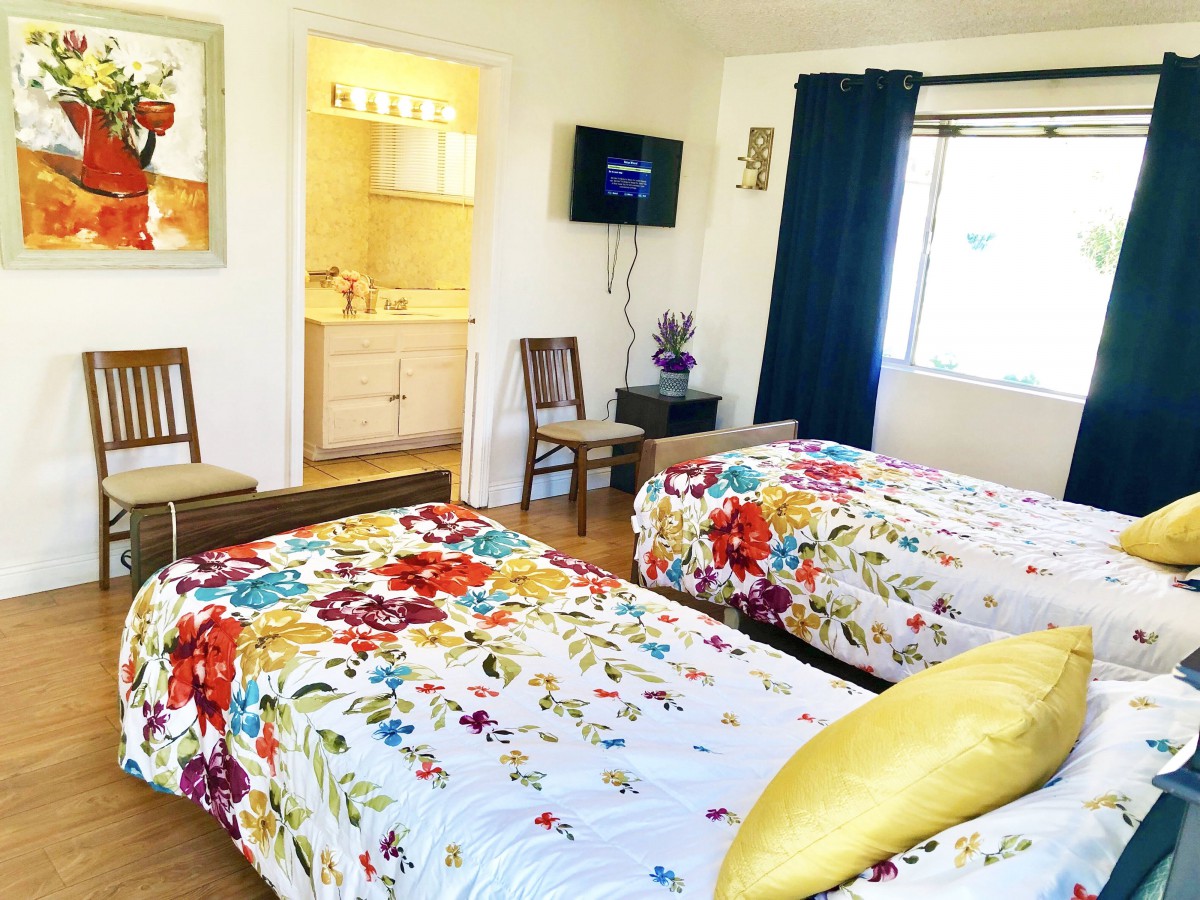 A bright bedroom featuring twin beds with colorful floral bedding, a wall-mounted TV, and a small bathroom in the corner.