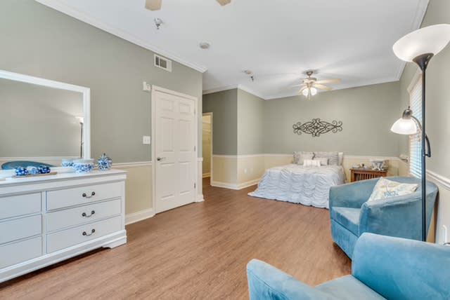 A spacious and elegantly decorated bedroom featuring light-colored walls, a comfortable bed, armchairs, a dresser, and hardwood flooring.