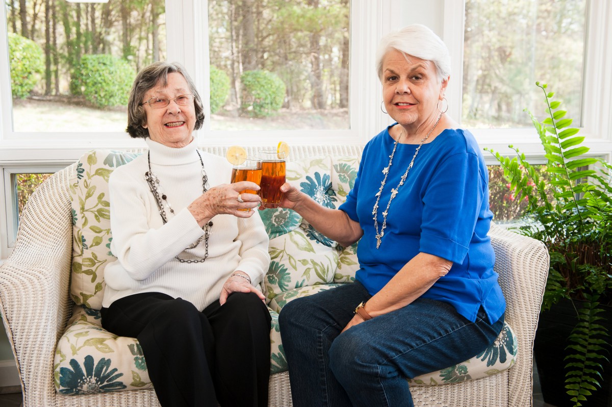 Two elderly women toasting with drinks while sitting on a couch indoors.
