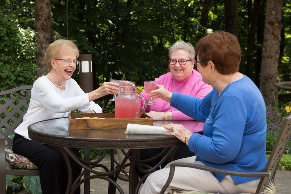 Three elderly women enjoying drinks and laughter together outdoors.