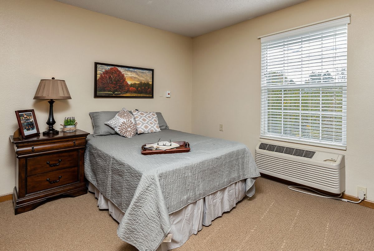A cozy bedroom with a neatly made bed, bedside table, lamp, wall artwork, and a window with blinds.