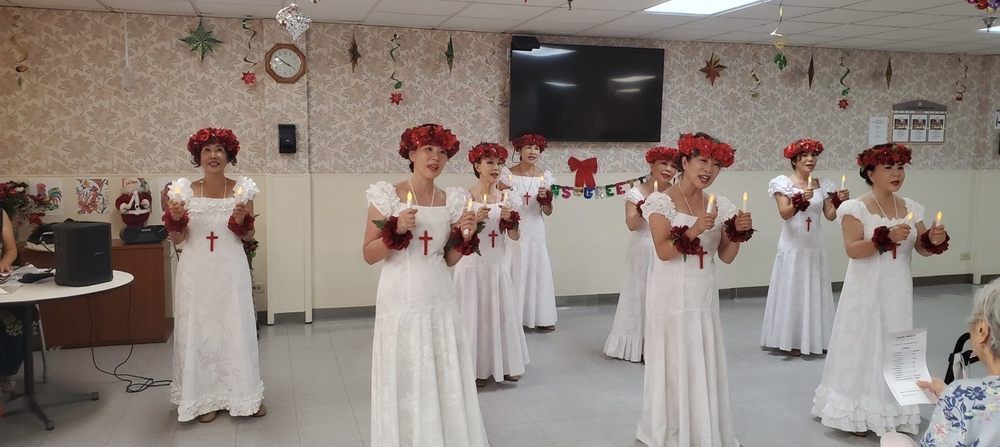 A group of women dressed in white gowns with red flower crowns, holding candles during an indoor performance.
