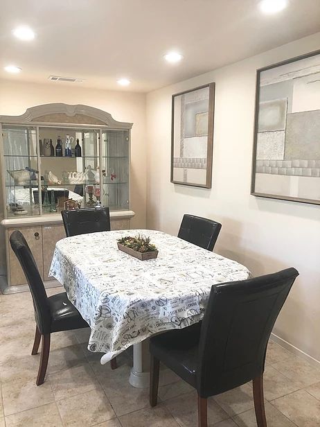 A cozy dining room featuring a table covered with a patterned cloth, surrounded by dark chairs. A cabinet with decorative items is in the background, and framed artwork adorns the walls.