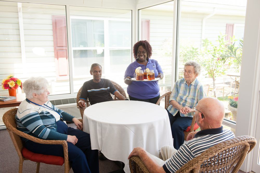 A caregiver serves drinks to a group of elderly individuals sitting around a table.