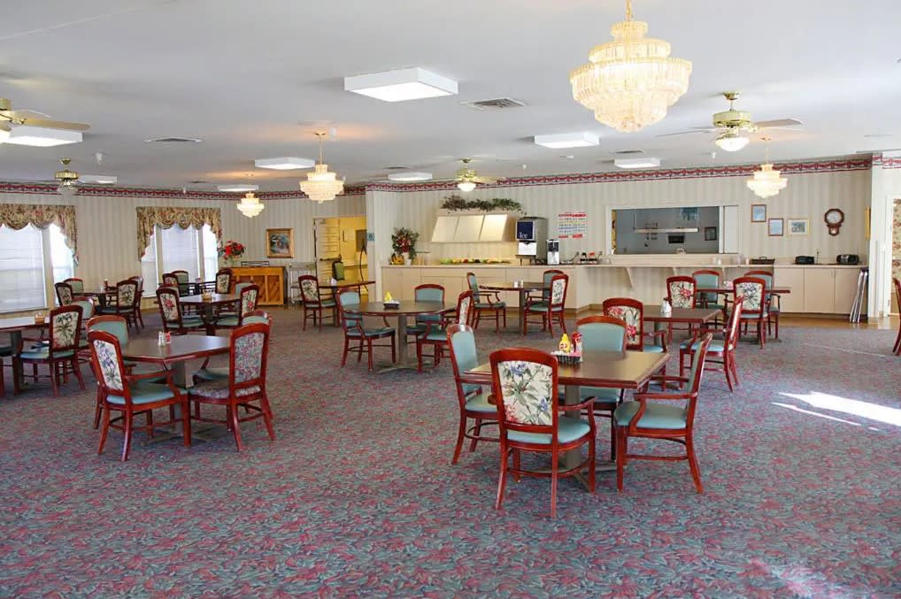 A spacious dining room with multiple round tables and chairs, featuring chandeliers and carpeted flooring.