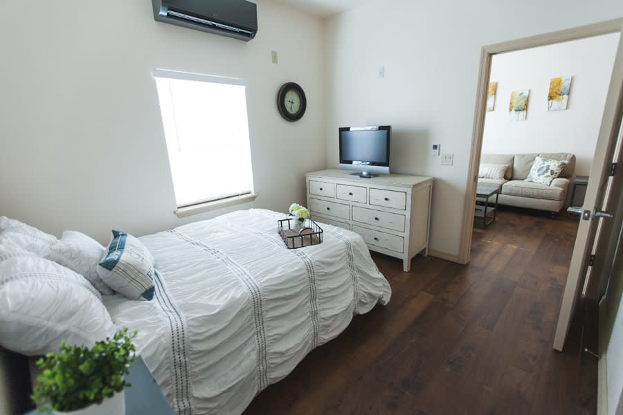 A minimalist bedroom with a white bed, dresser, and a TV, featuring wood flooring and natural light.