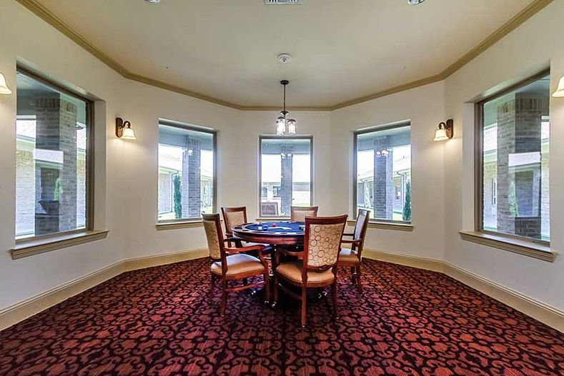 A cozy dining area featuring a round table with four chairs, surrounded by large windows and illuminated by a central light fixture, all set on a patterned carpet.