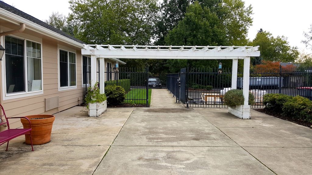 A driveway leads to a gated area under a white pergola, with potted plants on either side.
