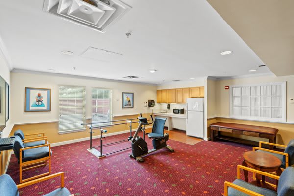 A therapy room with exercise equipment and a small kitchen area, featuring a carpeted floor.