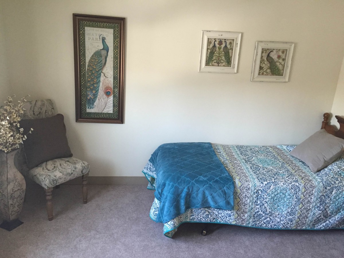 A cozy bedroom with a single bed covered in a blue blanket, adorned by artwork including a peacock painting and framed pictures on the wall.