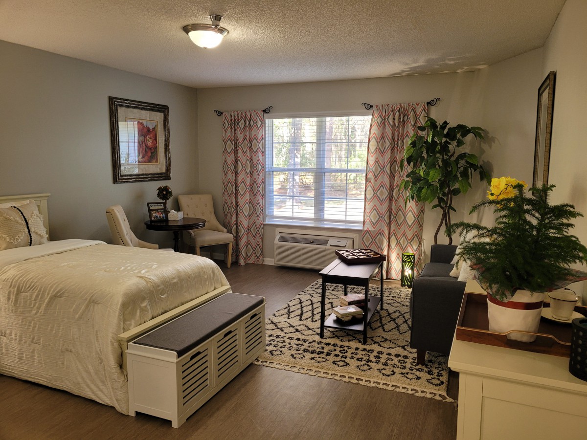 A cozy bedroom interior with a bed, seating area, and a large window letting in natural light.