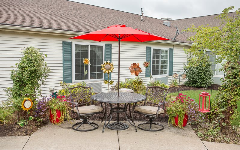 A cozy patio setting featuring a round table with four chairs and a vibrant red umbrella, surrounded by potted plants in front of a house.