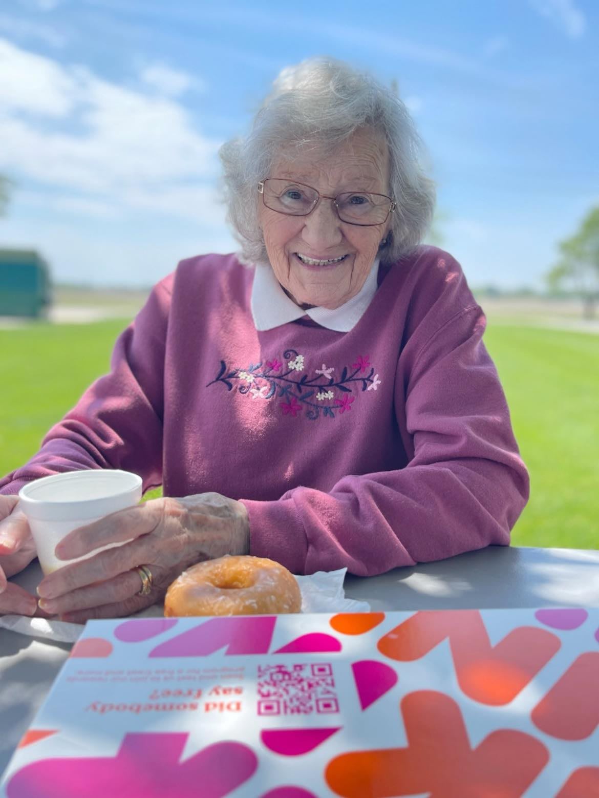 An elderly woman enjoying a donut and coffee at a picnic table outdoors.