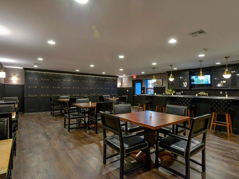 A modern restaurant interior featuring wooden tables and chairs with a bar area in the background.