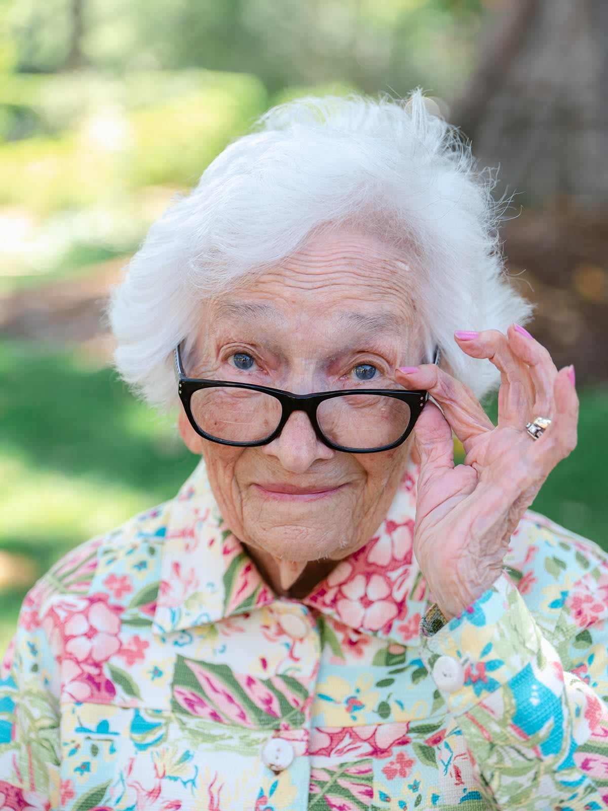 An elderly woman wearing glasses and a floral shirt smiles outdoors.