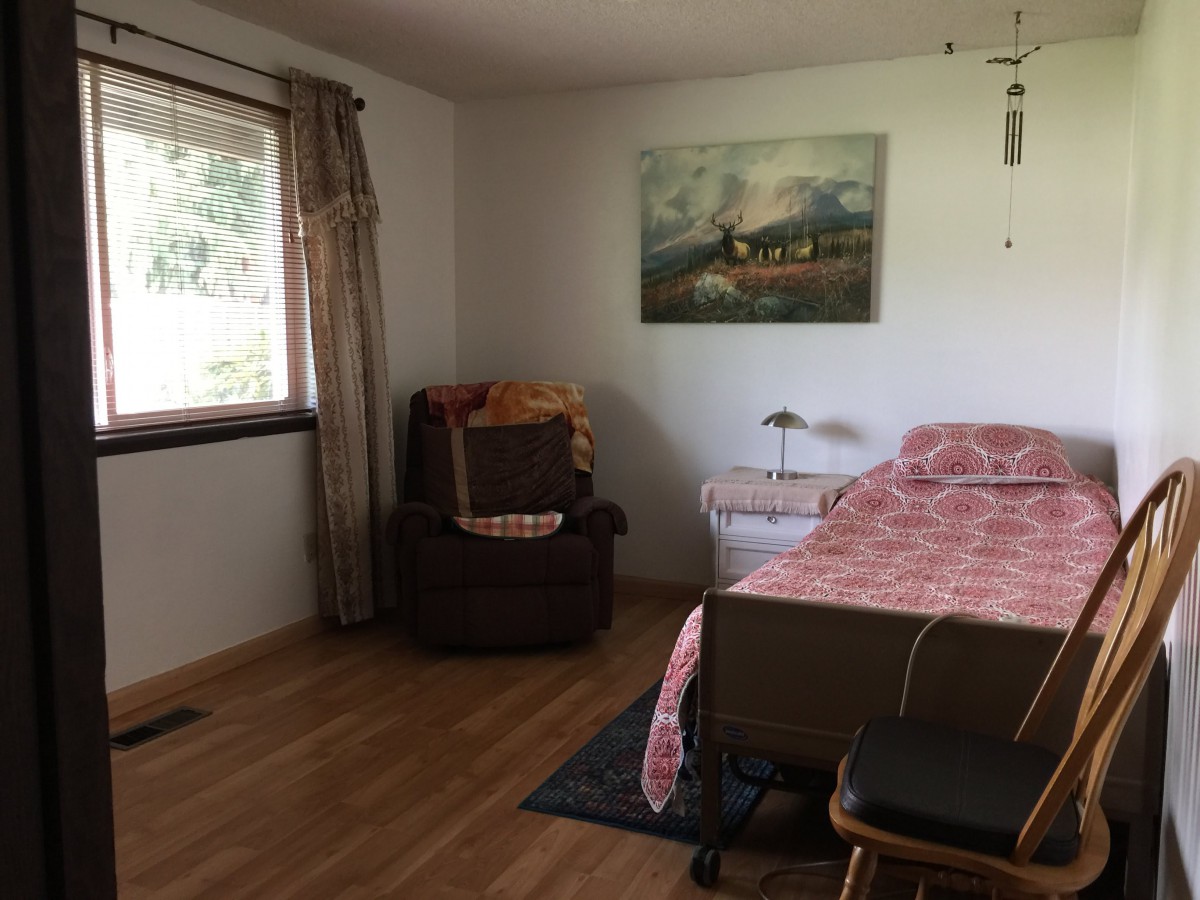 A cozy bedroom with a bed, chair, and painting on the wall, featuring wooden flooring and a window with curtains.