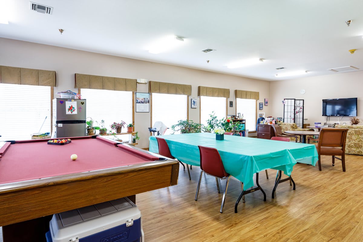 A spacious recreation room featuring a billiards table and tables with chairs, decorated with plants.