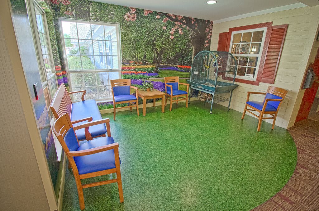 A brightly decorated waiting room with a green floor and colorful mural on the wall, featuring several wooden chairs and a table.