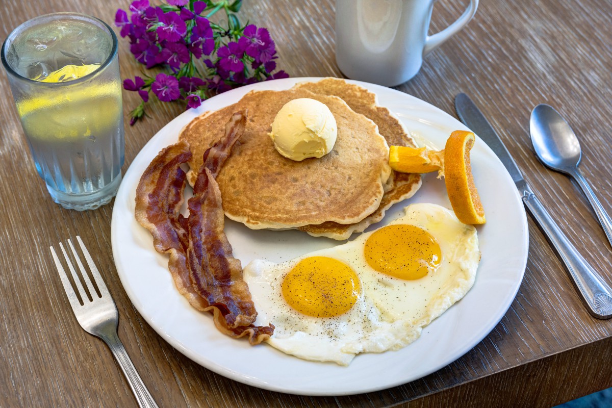 A hearty breakfast plate featuring pancakes with butter, fried eggs, crispy bacon, and a slice of orange, accompanied by a cup of coffee and flowers in the background.