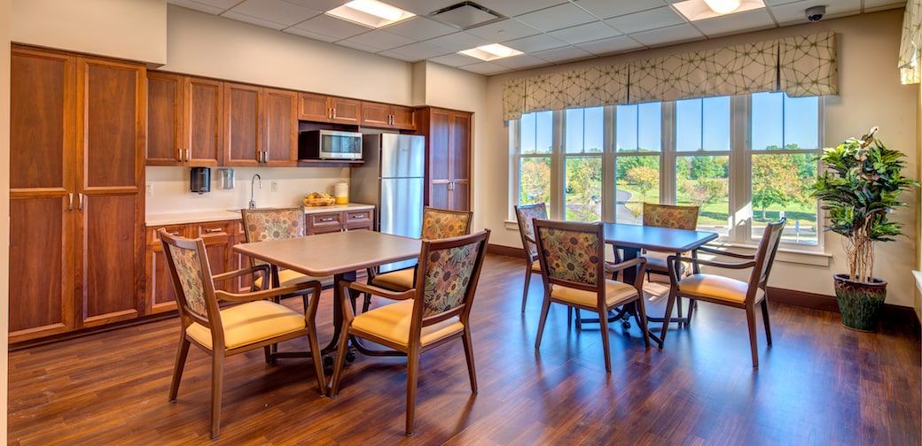 A cozy kitchen and dining area with wooden floors, featuring a fridge, microwave, and tables with chairs by the window.