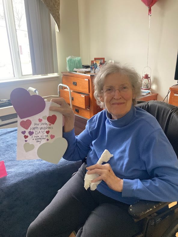 An elderly woman smiles while holding a heart-themed card indoors.