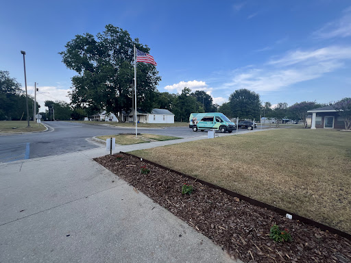 A view of a park area featuring a flag, trees, and parked vehicles under a clear blue sky.