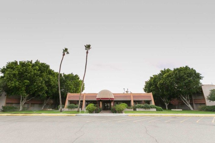 A single-story building with a red-tiled roof and an entrance surrounded by palm trees against an overcast sky.