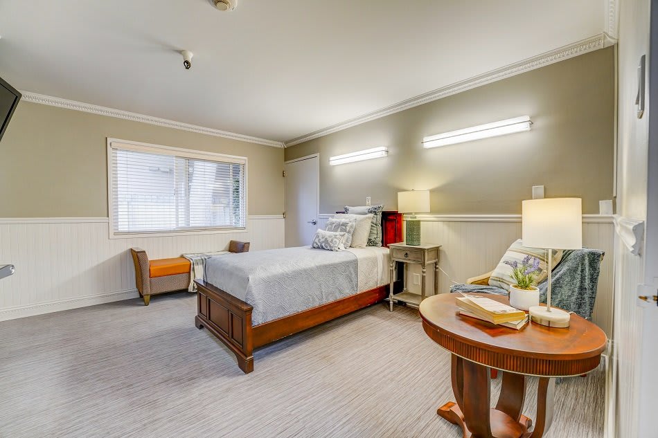 A cozy bedroom with a neatly made bed, bedside table and lamp, window, and additional seating.