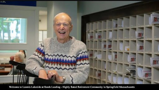 An elderly man smiles warmly while standing in front of mailboxes at a community center.