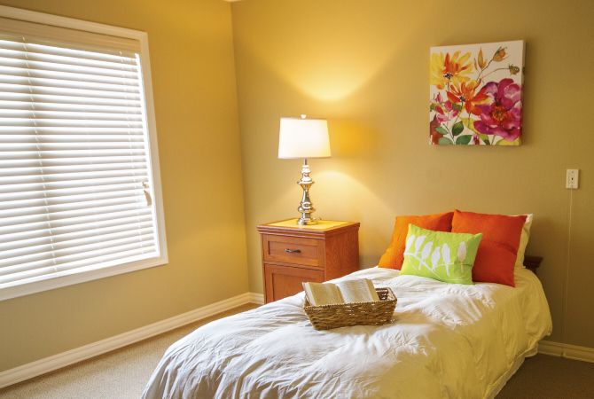 A cozy bedroom with a neatly made bed, colorful pillows, a nightstand with a lamp, and a decorative wall painting.