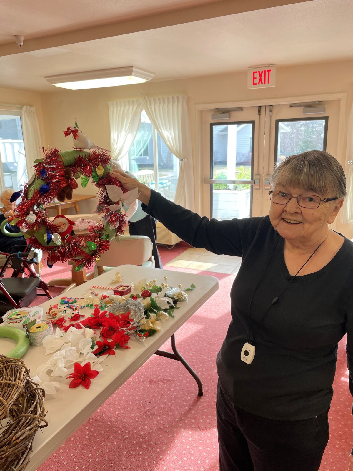 A woman happily displaying a handcrafted wreath in an indoor setting with various decorations on a table.