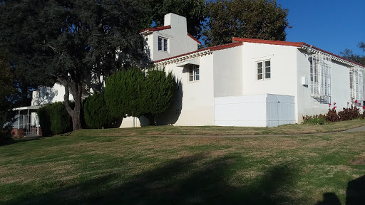 A white residential building with a red tiled roof, surrounded by green grass and trees.