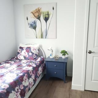A minimalist bedroom with a single bed featuring floral bedding, a nightstand with a lamp and plant, and vibrant artwork on the wall.