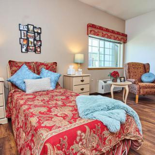 A cozy bedroom with a single bed featuring red and gold bedding, a blue throw blanket, and decorative pillows. The room includes a nightstand with a lamp, an armchair, and framed photographs on the wall.