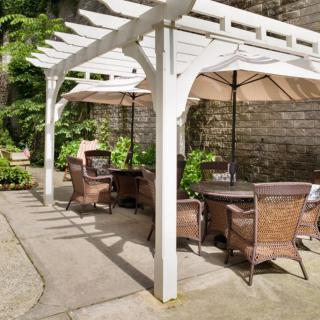 A cozy outdoor patio area with wicker seating under a white pergola, complemented by large umbrellas and surrounded by a garden with a stone wall backdrop.