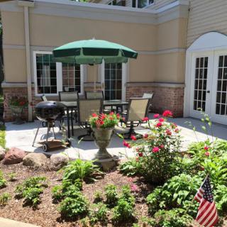 A sunny outdoor patio space featuring a dining set, grill, and a vibrant garden.