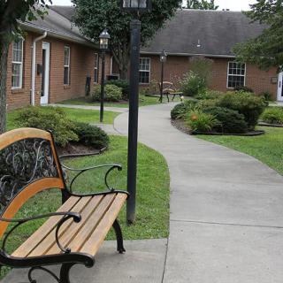 A peaceful garden setting with a bench beside a concrete pathway leading to brick buildings.