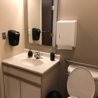A clean and accessible bathroom with a sink, mirror, toilet, and dispensers.